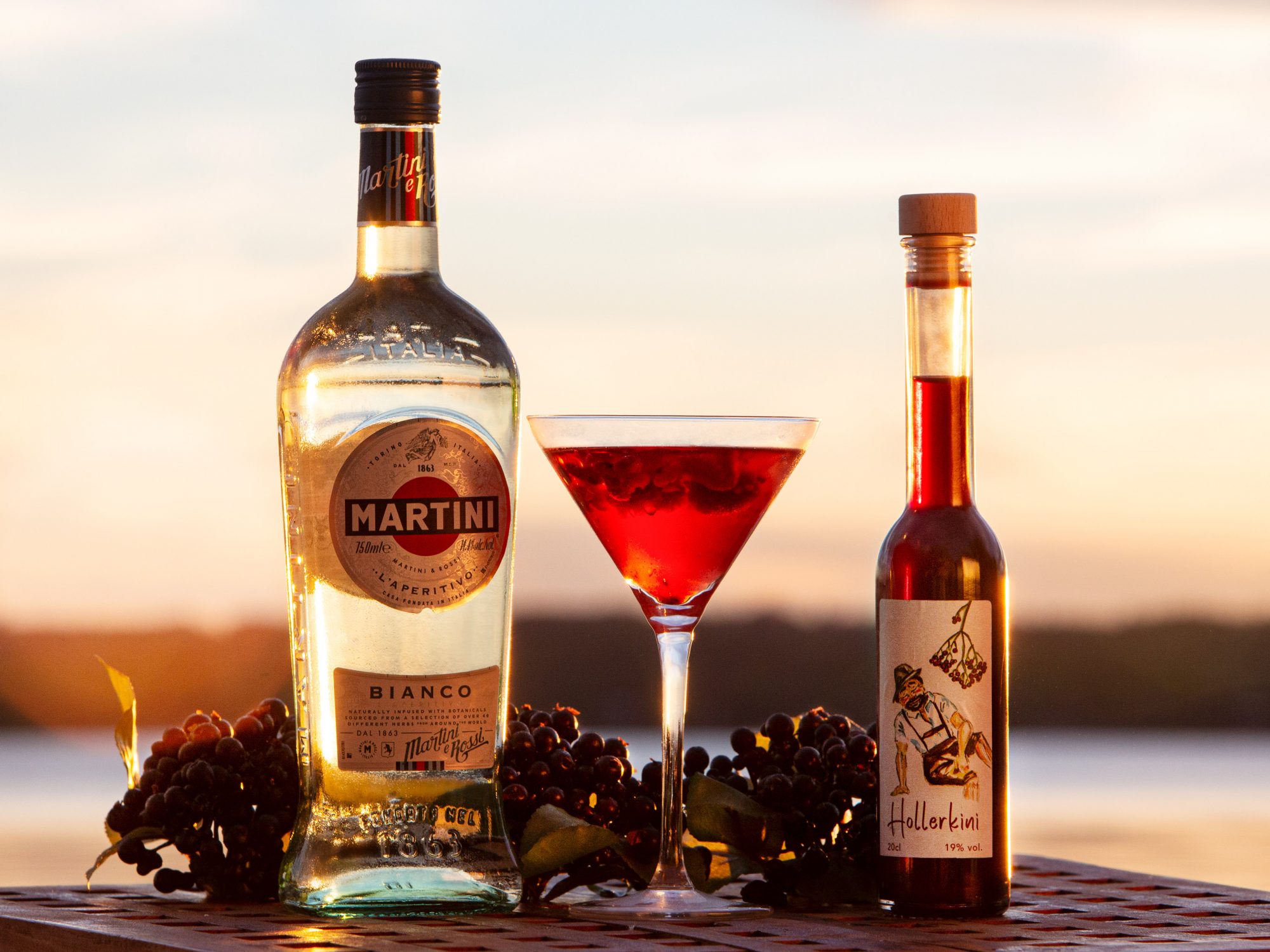 Eine Flascche Sekt links, eine Flasche Hollerkini rechts und ein Daikini im Martiniglaß in der Mitte auf einem Holztisch vo dem Sonnenuntergang beim Ammersee.