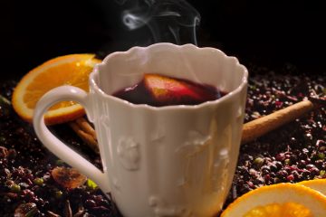 Heißer Holler (Apfelsaft und Hollerkini erhitzt) in einer weißen Tasse vor schwarzem Hintergrund mit Orangen, Zimtstangen und Holunderbeeren ringsherum.