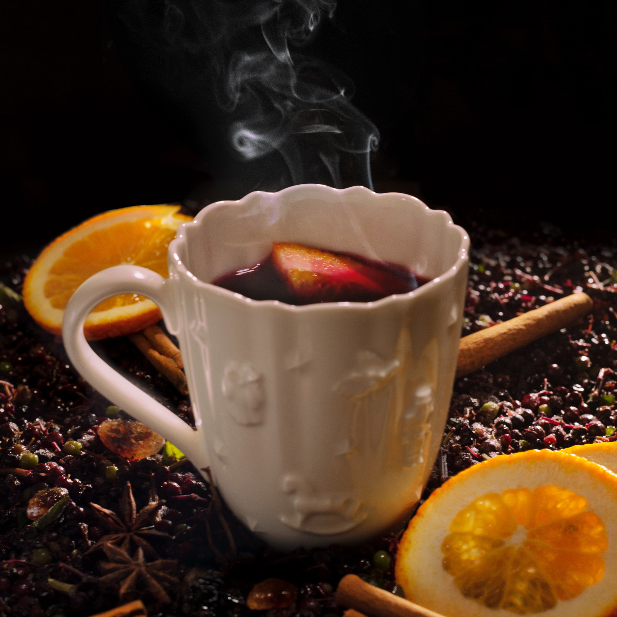 Heißer Holler (Apfelsaft und Hollerkini erhitzt) in einer weißen Tasse vor schwarzem Hintergrund mit Orangen, Zimtstangen und Holunderbeeren ringsherum.